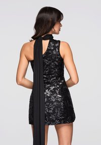 Svart ärmlös klänning med blommig paljettbroderi, hög halslinning och en lång, flytande tygsjal knuten baktill. Kort fåll.