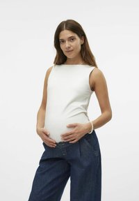 Top blanc sans manches avec un col montant, associé à un jean en denim bleu ample. Le modèle caresse doucement un ventre arrondi visible.