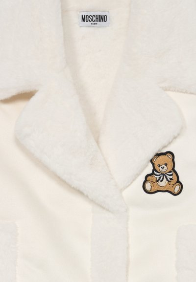 Cremfarbener Fleece-Jacke mit plüschiger Textur, ausgestattet mit einer schwarzen und braunen Teddy-Bären-Stickerei und einem klassischen Kragendesign.
