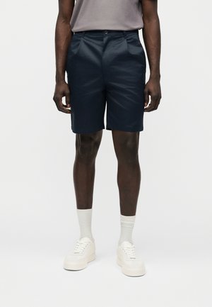 Muž nosi tmavomodré na mieru šité šortky, svetlosivé tričko, biele ponožky a biele tenisky, stojaci na jednoduchom pozadí.