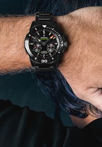 Melna metāla pulksteņa apaļa sejas daļa ar kontrastējošām baltām un zaļām atzīmēm, datuma logu un teksturētu melnu siksniņu, kurš valkāts uz rokas.