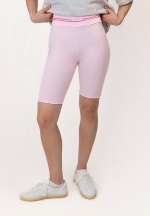 Personne portant un short de cycliste rose clair avec une ceinture rose et des baskets blanches, debout, les mains tirant légèrement un t-shirt gris vers le haut.