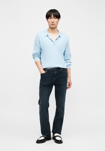 Jeune homme debout, portant un polo à manches longues bleu clair, un jean foncé et des mocassins noirs et blancs, devant un fond uni clair.