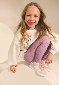 Jasnoróżowe sneakersy z białymi paskami, lawendowe legginsy z wzorem w formie linii oraz kremowy sweter z dekoracyjnymi akcentami.