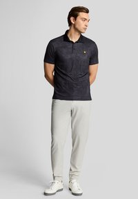 Sort polo-shirt med et diskret mønster og et guld logo på brystet, parret med lysegrå bukser og hvide sneakers.
