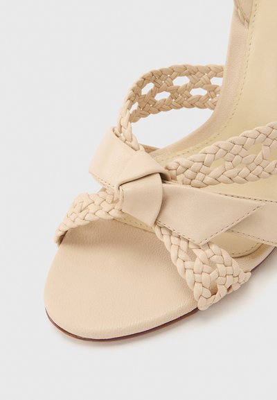 Μπεζ σανδάλι με πλεκτούς δερμάτινους ιμάντες και λεπτομέρεια με δερμάτινο κόμπο πάνω από το ανοιχτό δάχτυλο και την περιοχή του αστραγάλου.