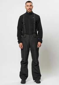 Mustat vedenpitävät haalari, joissa on säädettävät hihnat, yhdistettynä mustaan vetoketjulliseen fleece-takkiin. Fleece-takissa on logo ja haalareissa valkoinen korostus.