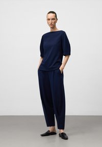 Vrouw die staat met haar handen in de zakken, gekleed in een marineblauwe top met mouwen tot de ellebogen, bijpassende wijde broek en zwarte instapschoenen.