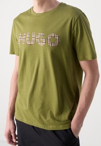 Olivewą zieloną koszulkę bawełnianą z krótkim rękawem, z okrągłym dekoltem, z teksturowanym logo "HUGO" w beżowym i czarnym kolorze, w kratkę houndstooth, umieszczonym na piersi.