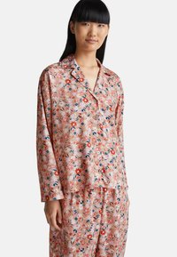 Donna che indossa una camicia da pigiama rosa floreale con bottoni e pantaloni abbinati, in piedi su uno sfondo bianco semplice.