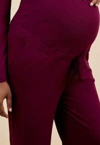 Pantalons de maternité en tissu bordeaux foncé, dotés d'une taille à cordon, de poches latérales et d'une texture lisse. S'adapte à un ventre arrondi.