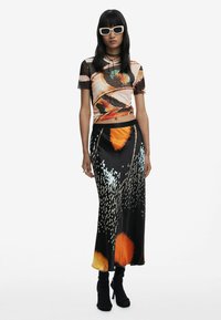 Le mannequin porte un haut transparent ajusté avec un motif de papillons, associé à une longue jupe noire aux accents colorés et à la texture lisse.