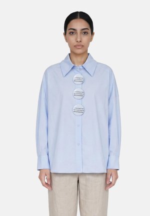 Femme aux cheveux foncés porte une chemise bleu clair oversize avec de gros boutons ronds affichant des chiffres, associée à un pantalon beige sur fond blanc.
