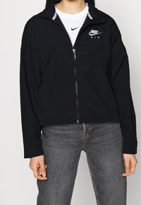 Svart kort jacka med dragkedja fram, med hög krage, två framfickor och en Nike Air-logga på bröstet. Tillverkad av lättviktig tyg.