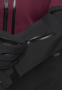 Schwarze wasserdichte Jacke mit oberem Abschnitt in Kastanienbraun, ausgestattet mit einer Reißverschlusstasche am Ärmel und strukturierten Handschuhen, die den Stoff greifen.