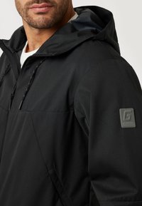 Juodas gobtuvas striukė su lygiu tekstūros paviršiumi, pasižyminti užtrauktuku užsegama kišene, virvutėmis ir prekės ženklo emblema ant rankovės.