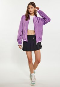 Paarse bomberjack met witte ribgebreide manchetjes, gedragen over een cropped wit topje, gecombineerd met een zwarte plooienrok en meerkleurige sneakers.
