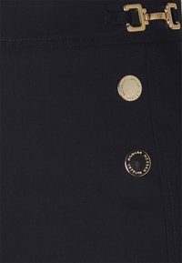 Primer plano de tela negra con dos botones redondos dorados grabados con "Morgan" y un broche metálico con hebillas rectangulares doradas.