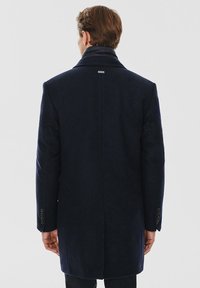 Granatowy wełniany płaszcz o fasonie strukturalnym, z wysokim kołnierzem i mankietami na guziki. Charakterystyczny detal z metką na plecach.