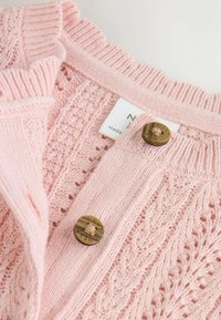 Cardigan rosa lavorato a maglia con motivo a traforo, polsini a costine e due bottoni tondi marroni. Presenta un'etichetta con la scritta "NUOVO". Texture morbida.