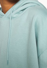 Sudadera azul claro con un bolsillo tipo canguro en la parte frontal, capucha con cordones y puños acanalados. Textura de tela suave con un diseño simple y relajado.