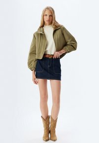 Oliven grøn cropped jakke med elastiske manchetter, creme top, denim miniskørt, brun bælte med en cirkulær spænde, og brune cowboy støvler.