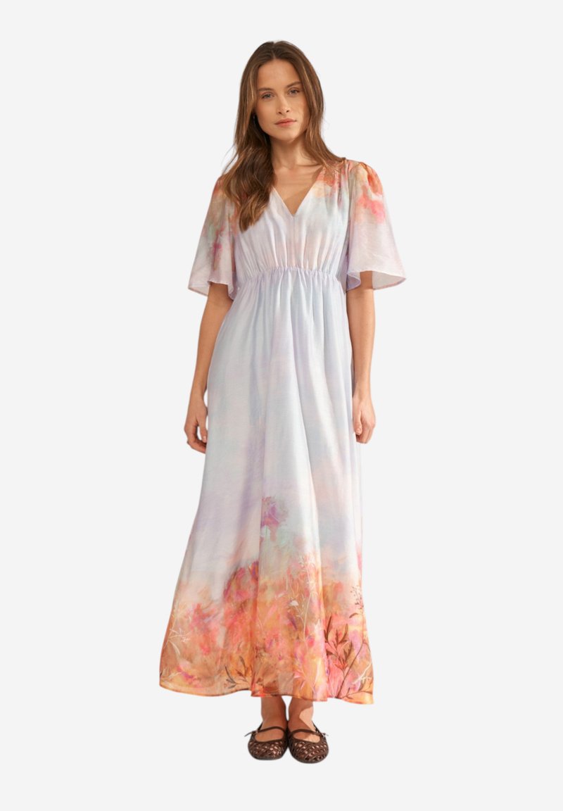 Mujer de pie con un vestido largo hasta el suelo, de flores en tonos pastel, manga corta y bailarinas negras, sobre un fondo blanco liso.