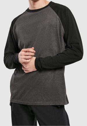 Hombre con camisa de manga larga gris oscuro y mangas negras, con las manos entrelazadas a la altura de la cintura, sobre un fondo claro y uniforme.
