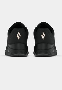 Paire de chaussures de sport noires vues de dos avec des talons relevés et un logo de couleur claire en forme de losange sur chaque talon.