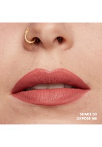Rossetto opaco in una delicata tonalità pesca rosa, texture morbida, evidenzia labbra piene, con un anello nasale dorato come accento. Il testo dice "SHADE 03 XXPOSE ME."