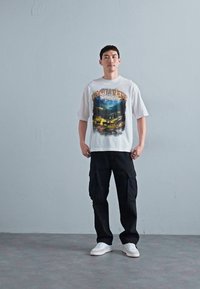 Mand står med ryggen mod en grå væg iført en hvid grafisk t-shirt, sorte cargo bukser og hvide sneakers, vendt fremad med hænderne langs siden.