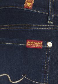 Vaqueros de mezclilla azul oscuro con una etiqueta roja con letras doradas, costuras doradas y un detalle distintivo de bordado curvado blanco en el bolsillo.