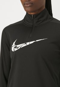 Svart långärmad kvartdraget topp med en texturerad vit Nike-logotyp över bröstet. Tyget ser jämnt och åtsittande ut, med en hög krage.