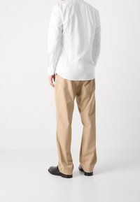 Bílá košile s dlouhým rukávem a hladkou texturou, kombinovaná s béžovými kalhotami. Černé boty dotvářejí outfit. Pohled ze zad, minimalistické pozadí.