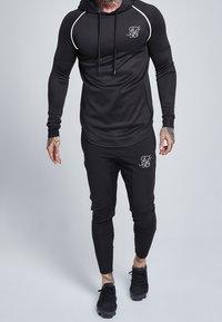 Schwarzes Sport-Outfit mit einem langärmeligen, kapuzenförmigen Oberteil mit weißen Akzenten und passenden, schmal geschnittenen Hosen mit Logodetails. Texturiertstoff.