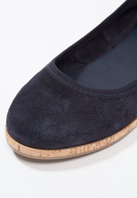 Gros plan d'une chaussure plate en daim bleu foncé avec un bout arrondi et une semelle claire à texture liège sur fond blanc.