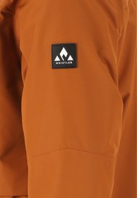 Orangene wasserdichte Jackenmaterial mit glatter Textur. Verfügt über einen schwarzen quadratischen Aufnäher mit dem „WHISTLER“-Logo und einem Bergdesign.