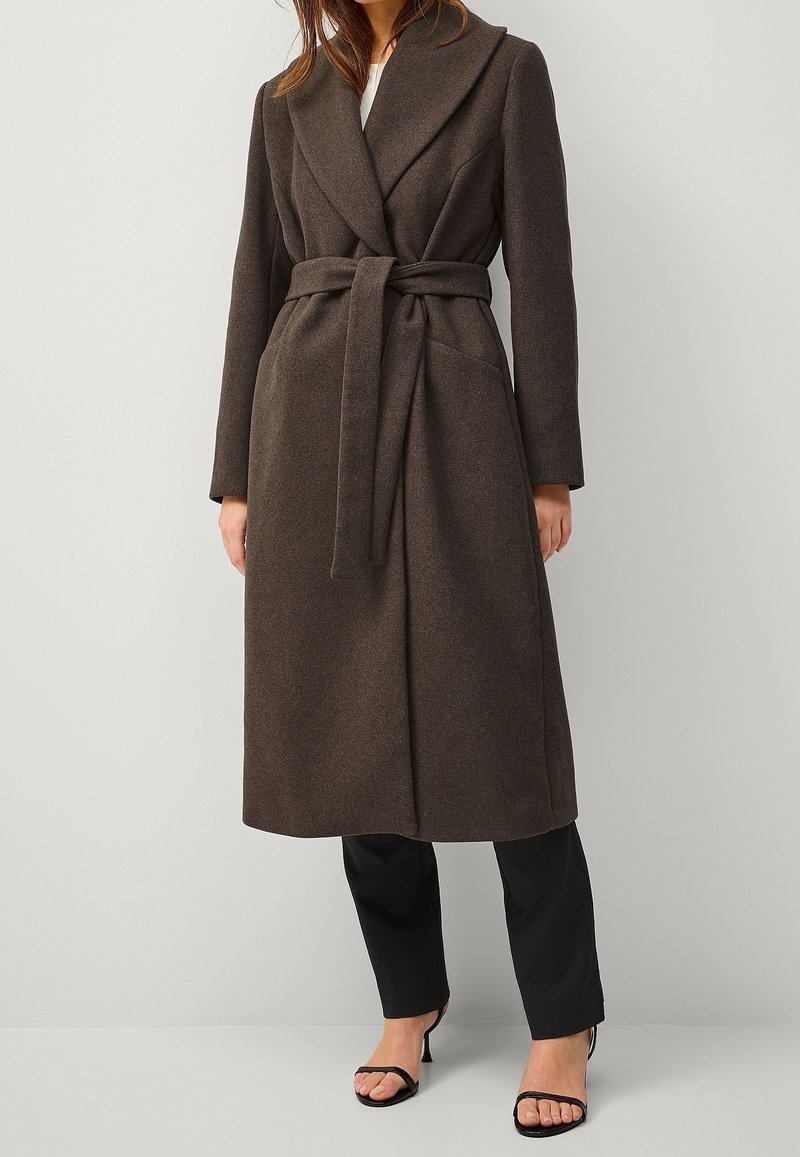 Femme portant un long manteau marron foncé ceinturé sur un pantalon noir et des sandales noires à talons, debout devant un fond uni et clair.