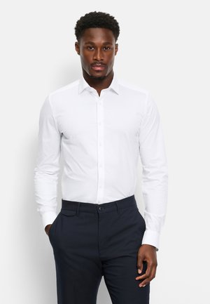 Man wearing a fitted white dress shirt and dark navy trousers, standing with hands partially in pockets against a plain background.