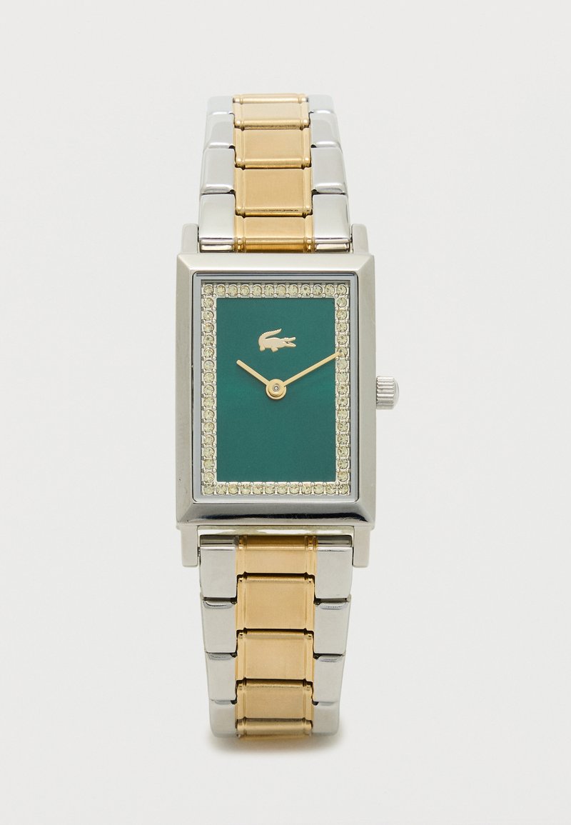 Klaasi kujuga kell, mille teal värviline kellanägu on raamitud selgete kristallidega. Kellal on hõbedane ja kuldne roostevabast terasest rihm, ning selles on kuldne logo ja lihtsad käed.