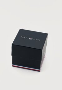 Μαύρο κουτί ρολογιού Tommy Hilfiger με λογότυπο στην κορυφή και κόκκινες, λευκές και μπλε λωρίδες κατά μήκος της κάτω άκρης σε λευκό φόντο.