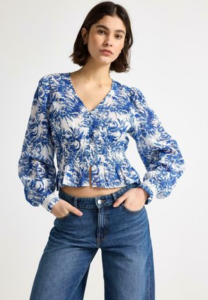 Donna con capelli corti e scuri che indossa una camicia a maniche lunghe blu e bianca con motivo floreale e jeans blu a vita alta con le mani nelle tasche.