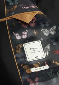 Sort stof med for i sommerfuglsmønster. Mærket læser "next tailoring," størrelse "40R," og "slank pasform" i hvid tekst. Gyldent kantbånd som accent.