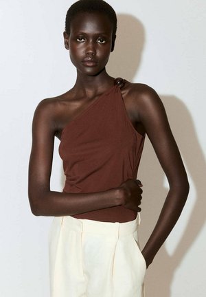 Femme à la peau foncée portant un haut marron sans manches avec une bretelle sur une épaule et un pantalon crème taille haute, debout devant un fond uni.