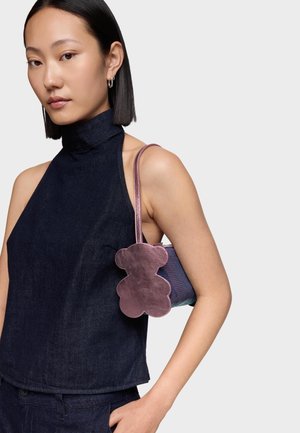 Mujer con cabello negro liso lleva un top oscuro sin mangas tipo halter y lleva un bolso pequeño con un detalle de oso de peluche rosa brillante en el hombro.