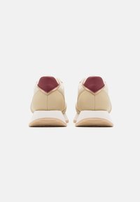 Sneaker beige con un accento maroon sul tallone, texture in pelle liscia e una suola bianca. Design semplice e sportivo con forma arrotondata.