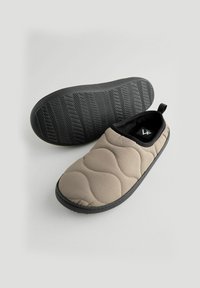 Zapatillas de casa acolchadas en color beige con una suela de goma negra texturizada, que cuentan con una punta redonda y una tira en el talón para facilitar su colocación.