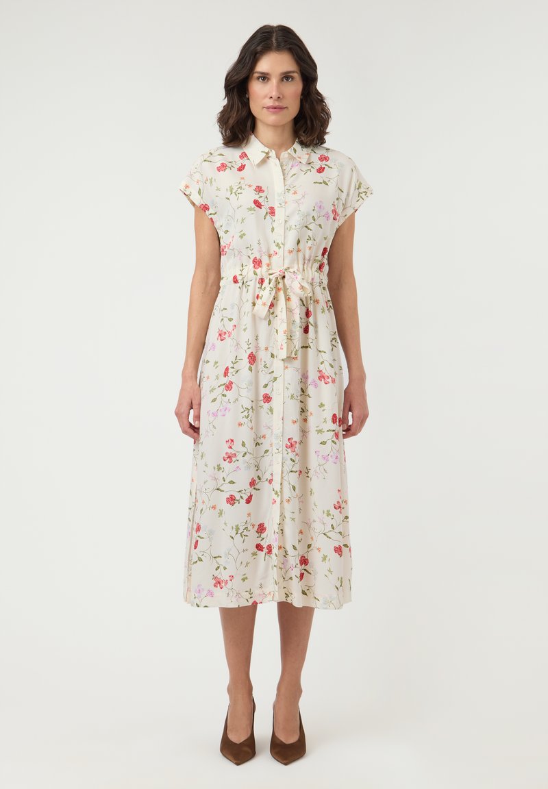 Γυναίκα που φορά άσπρο μίντι φόρεμα με ροζ και κόκκινο λουλουδάτο σχέδιο, κοντά μανίκια, κουμπιά μπροστά, ζώνη στη μέση και καφέ μυτερές γόβες.