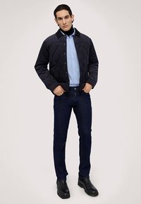 Chaqueta acolchada azul marino con cuello, usada sobre una camisa de rayas azul claro. Jeans oscuros ajustados y botas negras completan el conjunto.