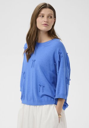 Žena oblečená v voľnom modrom svetri s malými mašličkami a bielej sukni, pozerajúca sa naľavo na jednoduchom pozadí.